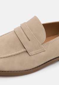 Mocassins en daim beige avec une semelle plate et un bout arrondi. Ils présentent une bande décorative et des détails de couture le long des bords.