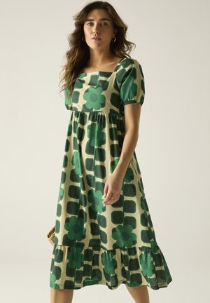 Vrouw die een beige midi-jurk draagt met groene geometrische en bloemmotieven, pofmouwen en een vierkante halslijn, staand tegen een effen achtergrond.
