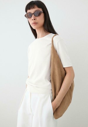 Femme portant une tenue blanche et des lunettes de soleil, portant un grand sac à bandoulière en daim beige, debout devant un fond clair uni.