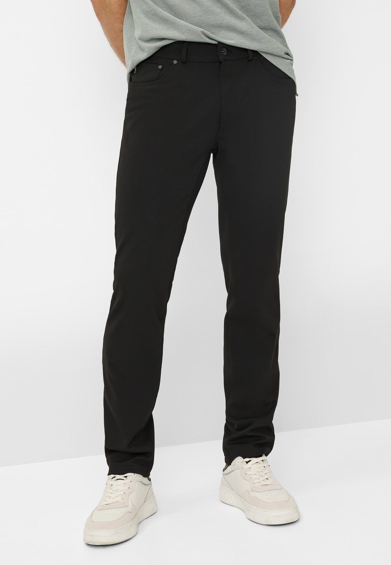 Chinos noirs cintrés avec une texture lisse, design à cinq poches, fermeture par bouton et accents métalliques discrets, associés à des sneakers claires.