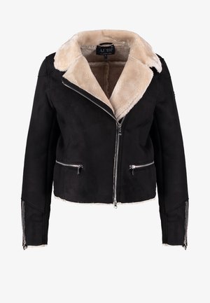 Vrouw draagt een zwarte schapenvachtjas, lichtroze blouse, zwarte skinny broek, zwarte enkellaarzen, houdt een zwarte handtas met studs vast.