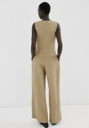 Persona in piedi di spalle che indossa una tuta smanicata beige con gambe larghe e mani nelle tasche su sfondo bianco.