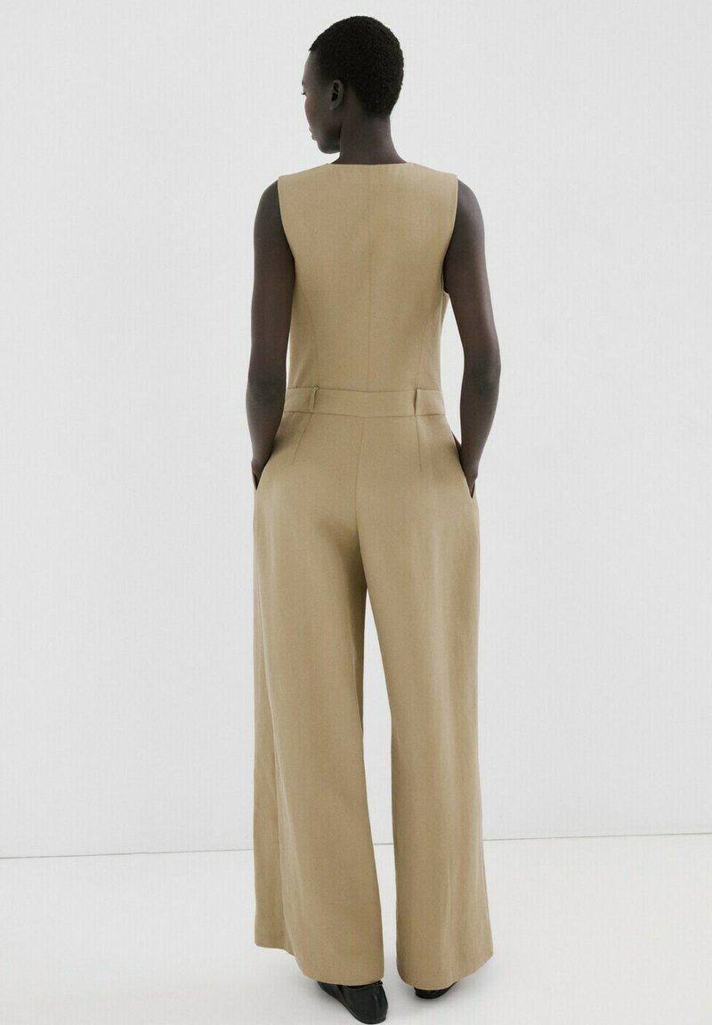 Osoba stojící zády k divákovi, oblečená v béžovém bezrukávém overal s širokými nohavicemi a rukama v kapsách na bílém pozadí.