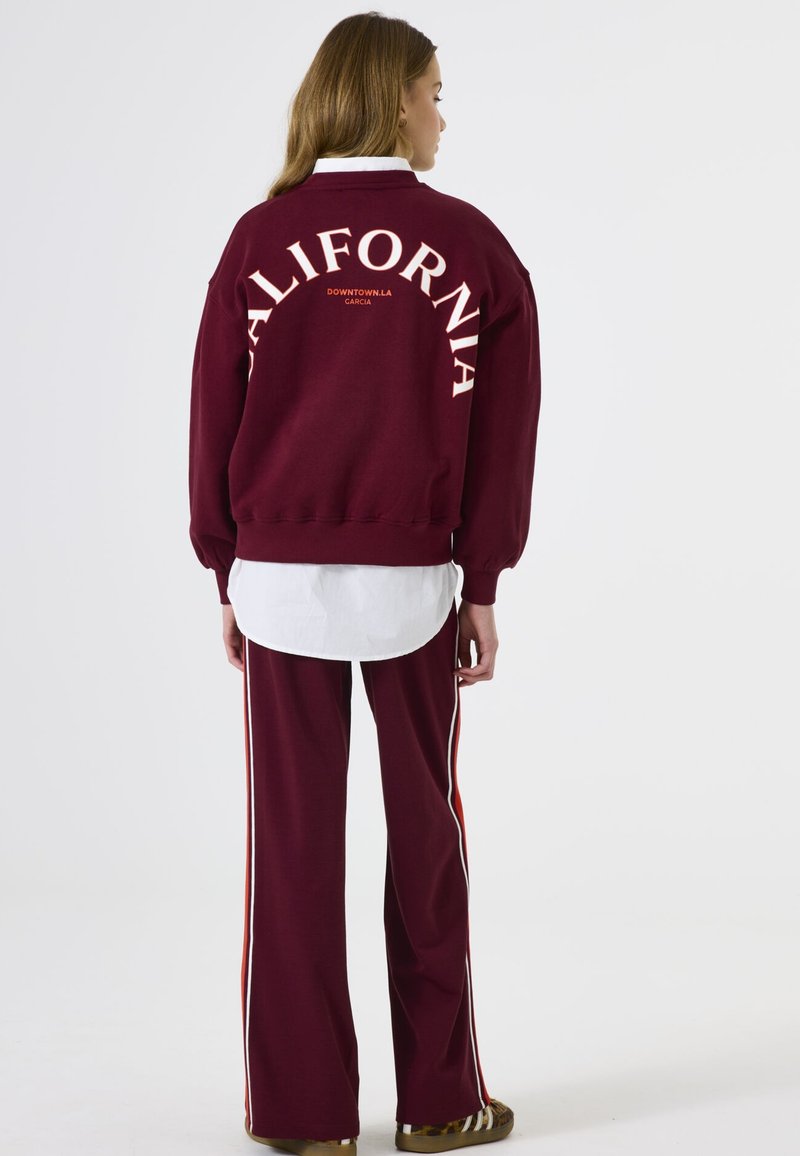 Sudadera burdeos con "CALIFORNIA" impreso en crema, combinada con pantalones burdeos a juego que tienen rayas laterales blancas y naranjas.