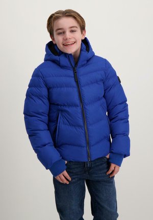 Smiling boy in a blue puffer jacket with a hood stands against a light background, hands in pockets, wearing jeans.