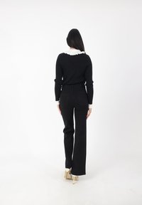 Suéter negro de canalé con un cuello blanco con volantes, combinado con jeans negros de cintura alta y zapatos de tacón de color claro. Vista trasera.
