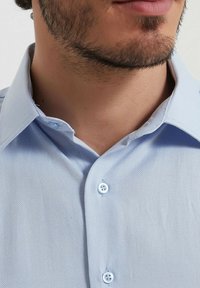 Chemise bleue claire à manches longues avec un tissu texturé, dotée d'un col classique et de deux boutons au niveau du cou, mettant en valeur de fines coutures.