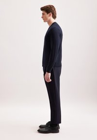 Cardigan a maglione di colore blu navy con scollo a V, maniche lunghe e chiusura con bottoni, abbinato a pantaloni scuri su misura e scarpe stringate nere.