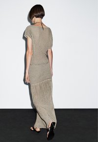 Gebreide jurk met beige strepen, korte mouwen, een losse pasvorm en zijsplitsingen. Gecombineerd met zwarte sandalen met bandjes. Achtergrond is effen wit.