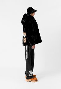 Chaqueta de piel sintética negra con capucha, que presenta un parche de oso. Combinada con pantalones holgados negros, zapatos de plataforma naranjas y un sombrero de cubo.