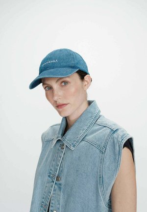Femme aux yeux bleus portant une veste sans manches en jean bleu et une casquette de baseball assortie sur un fond blanc uni.