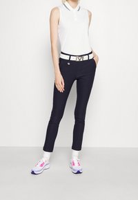 Polo bianco senza maniche con colletto, pantaloni attillati blu navy, cintura bianca con dettagli in argento e sneakers multicolori. Texture lisce ovunque.