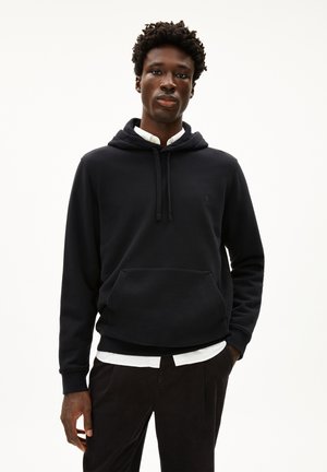 Black hooded sweatshirt made of soft fabric, featuring a kangaroo pocket, drawstrings, and a subtle logo on the chest. Paired with a white shirt.