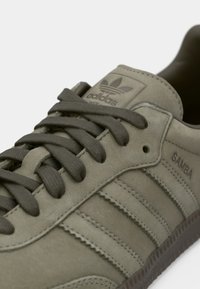 Sneakers z olivově zelené semiše s gumovou podrážkou, texturovanými tkaničkami a třemi bočními pruhy. Obsahují nápis „Samba“ a vystouplé logo Adidas.
