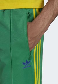 Hand i fickan på gröna Adidas träningsbyxor med gula ränder och blå Adidas-logga, kombinerade med en gul tröja.