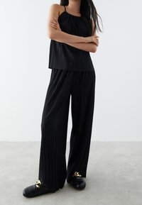 Svart plisserad linne med justerbara axelband som matchas med vid plisserad byxa. Har en slät yta och guld accenter på skorna.