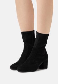Bottines en suède noir avec un bout rond, un talon épais et une fermeture éclair sur le côté. Elles présentent une texture douce et un design ajusté.