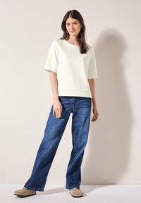 Top chiaro a maniche corte con testo in rilievo, abbinato a jeans blu a gamba larga e scarpe beige senza lacci su uno sfondo neutro.