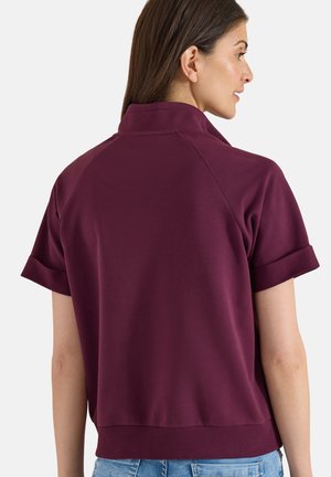 Kvinde med langt brunt hår iført en mørkelilla polo med korte ærmer og opfoldede kanter samt lyseblå jeans, set fra siden på en hvid baggrund.