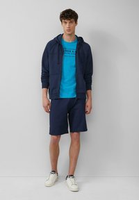 Marineblauer Zip-Hoodie über einem blauen T-Shirt mit grafischem Text, kombiniert mit marineblauen Shorts und weißen Sneakers. Legere Baumwollmischung.