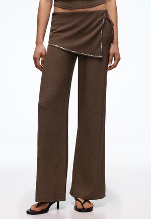 Brązowe spodnie z szerokimi nogawkami z asymetryczną cekinową nakładką w formie spódnicy, noszone z czarnymi sandałami na obcasie, na stojącej osobie.