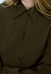 Chemise vert olive avec un col pointu, dotée d'une patte de boutonnage à l'avant et d'un détail de bouton unique. Tissu lisse avec une coupe structurée.