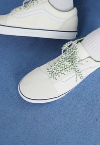 Crèmekleurige sneakers met witte accenten, groene geblokte veters en een gestructureerd oppervlak. Zwarte rand langs de zool, gezet op blauwe stof.
