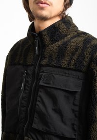 Fleece-takki tummanoliivinvihreänä ja mustana, jossa on pehmeä pinta, vetoketjullinen etuosa ja rintatasku, jossa on logo-patch.