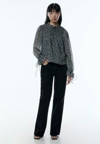 Zwarte en witte sheer blouse met bloemenpatroon, voorzien van een ruches halslijn en stropdasmanchetten, gecombineerd met rechte zwarte jeans en sandalen met banden.