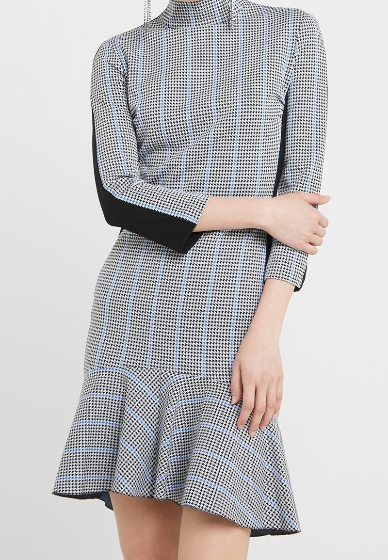 Femme portant une robe pied-de-poule noire et blanche avec des rayures bleu clair, des manches longues avec des panneaux noirs latéraux et un ourlet évasé.