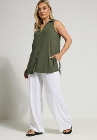 Tunika verde a costine senza maniche con colletto, chiusura a bottoni e tasche laterali, abbinata a pantaloni largo bianchi in lino e sandali.