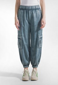 Jasnoniebieskie spodnie typu cargo z elastycznym pasem, mankietami przy kostkach i przednimi kieszeniami. Materiał ma miękką fakturę z wyblakłym wykończeniem.