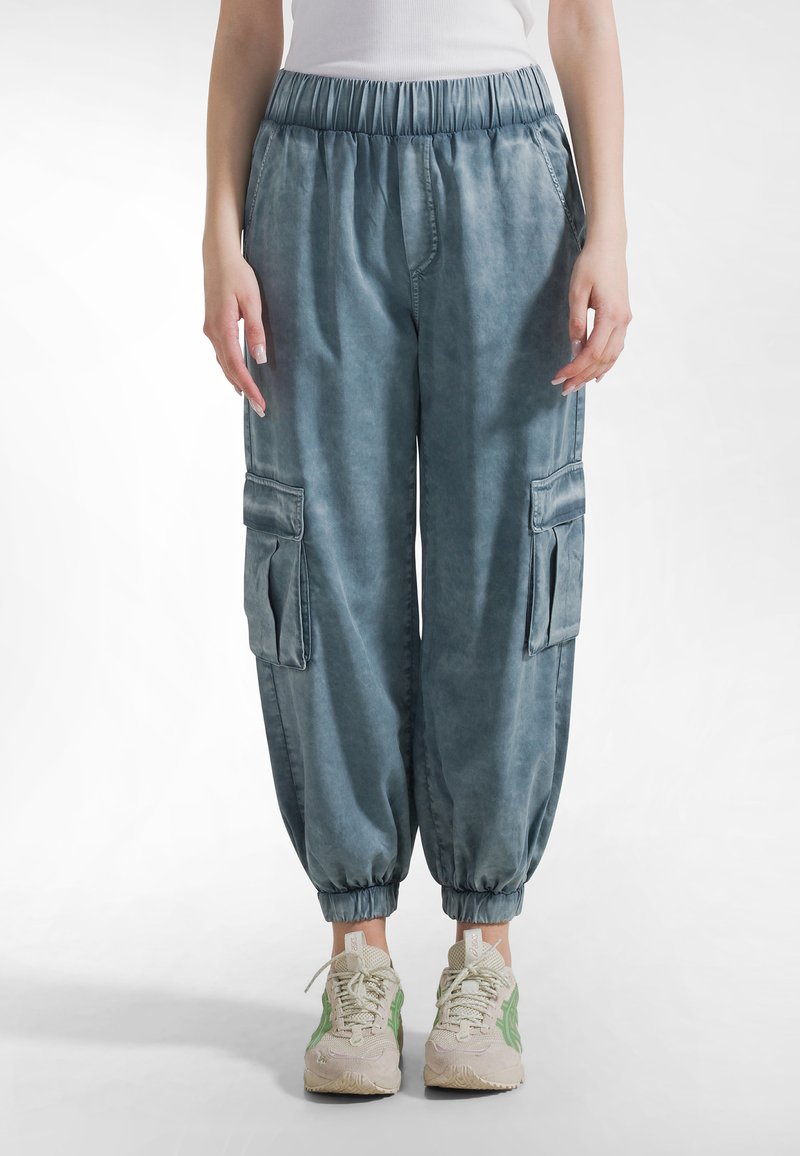 Jasnoniebieskie spodnie typu cargo z elastycznym pasem, mankietami przy kostkach i przednimi kieszeniami. Materiał ma miękką fakturę z wyblakłym wykończeniem.