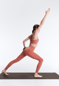 Ženska v aktivni obleki rjave barve drži utež za hrbtom, iztegne eno nogo naprej in nasprotno roko navzgor na črni blazini.