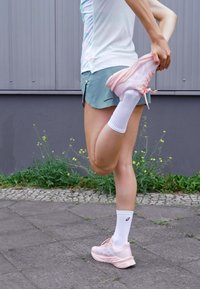 Light pink athletic shoes with a textured upper and contrasting laces, paired with white ankle socks. The person is stretching one leg.