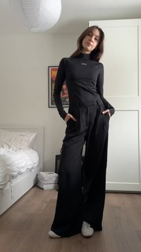 Mlada ženska stoji v spalnici, oblečena v črno dolgo majico z dolgimi rokavi in široke hlače, z rokami v žepih in belimi supergami.