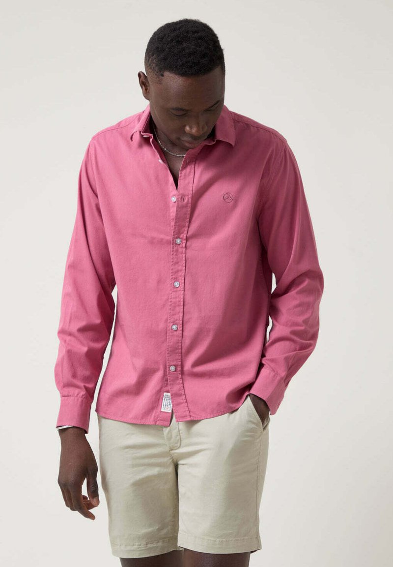 Camisa de botones de manga larga color rosa en tela ligera, con un pequeño logo en el pecho y un diseño de cuello clásico. Combinada con pantalones cortos beige.