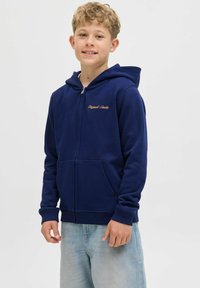 Pojke med ljust lockigt hår som bär en marinblå zip-up hoodie och ljusblå jeans, stående mot en enkel vit bakgrund.