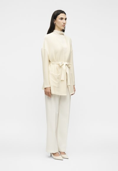 Γυναίκα που φοράει κρεμ ζακέτα με ζώνη και τσέπες, παντελόνια με φαρδιά πόδια και μυτερά τακούνια, στέκεται μπροστά σε ένα απλό λευκό φόντο.