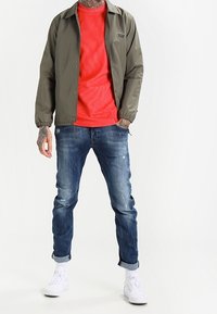 Oliivinvihreä takki, jossa on nepparit, punainen ribbipaita, siniset kuluneet farkut ja valkoiset lenkkarit. Kädet näkyvät tatuoinnit.