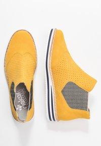 Żółte zamszowe botki za kostkę z perforowanym wzorem, elastycznymi bocznymi panelami i białą gumową podeszwą z granatowymi paskami oraz fakturowanym akcentem.