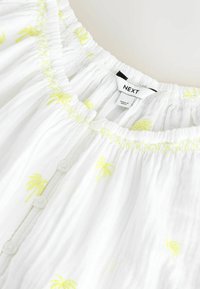 Vêtement blanc à boutons avec imprimé palmier jaune, encolure élastique avec surpiqûres jaunes et étiquette de marque "NEXT" cousue à l'intérieur du col.