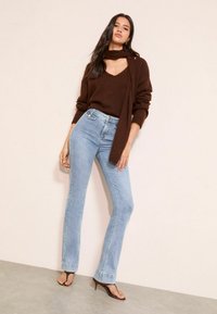 Hnedý pletený sveter s výstrihom do V a detailom šály, kombinovaný so svetlomodrými vysokými džínsami s rozšírenými nohavicami a čiernymi sandálmi na podpätku.