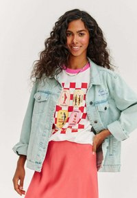 Jasnoniebieska kurtka dżinsowa z zapięciem na guziki, noszona na białej koszulce z kolorowymi grafikami ryb na czerwonej kratce, zestawiona z różową spódnicą.