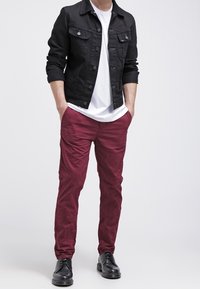 Veste en denim noir, t-shirt blanc, pantalon bordeaux et chaussures noires. La veste présente un design boutonné et des manches retroussées.