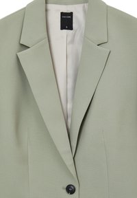 Hellgrüner Blazer mit Reverskragen und einzelner schwarzer Knopfverschluss. Glatte Textur, Innenfutter aus einem helleren Stoff. Markenschild sichtbar.