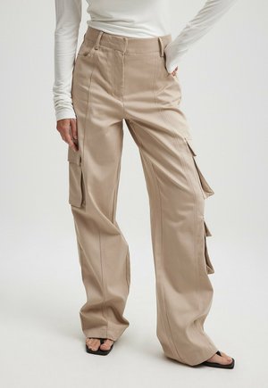 Pantaloni cargo beige realizzati in tessuto robusto con gambe larghe, dotati di tasche laterali e alette laterali, abbinati a sandali neri con fascia.