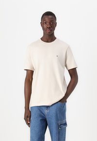 T-shirt beige à manches courtes en coton, doté d'un col rond et d'un petit emblème de marque sur la poitrine. Porté avec un jean cargo bleu.