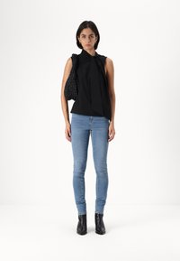 Top preto sem mangas com detalhe em laço, emparelhado com jeans azuis claros justos e botas de tornozelo pretas, acompanhado por uma bolsa preta texturizada.