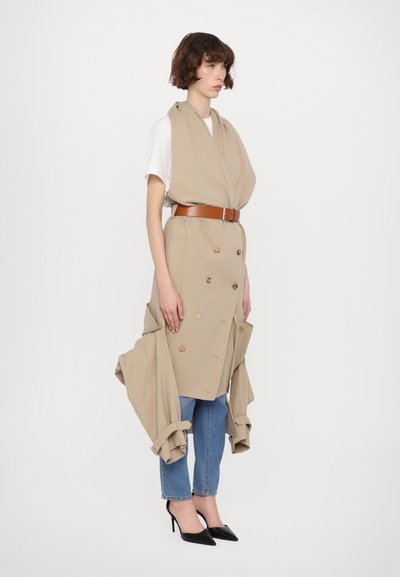 Beige, doppelt geknöpfter ärmelloser Mantel mit großen Knöpfen, braunem Gürtel, kombiniert mit einem weißen Hemd, blauen Jeans und schwarzen spitzen High Heels.
